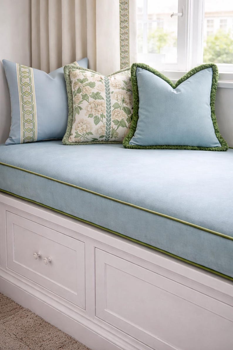 May include: A window seat with a light blue cushion and pillows. The pillows have decorative trim and patterns, including floral and geometric designs. The seat has a white storage base with two drawers.