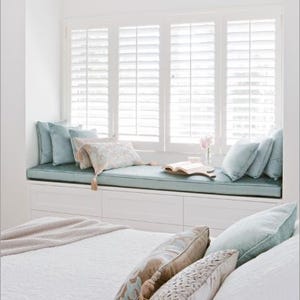 May include: A bright, airy bedroom with a window seat and bed. The window seat has a light blue cushion and pillows. The bed has white bedding and decorative pillows. A ceiling fan is visible. The room is decorated in neutral tones.
