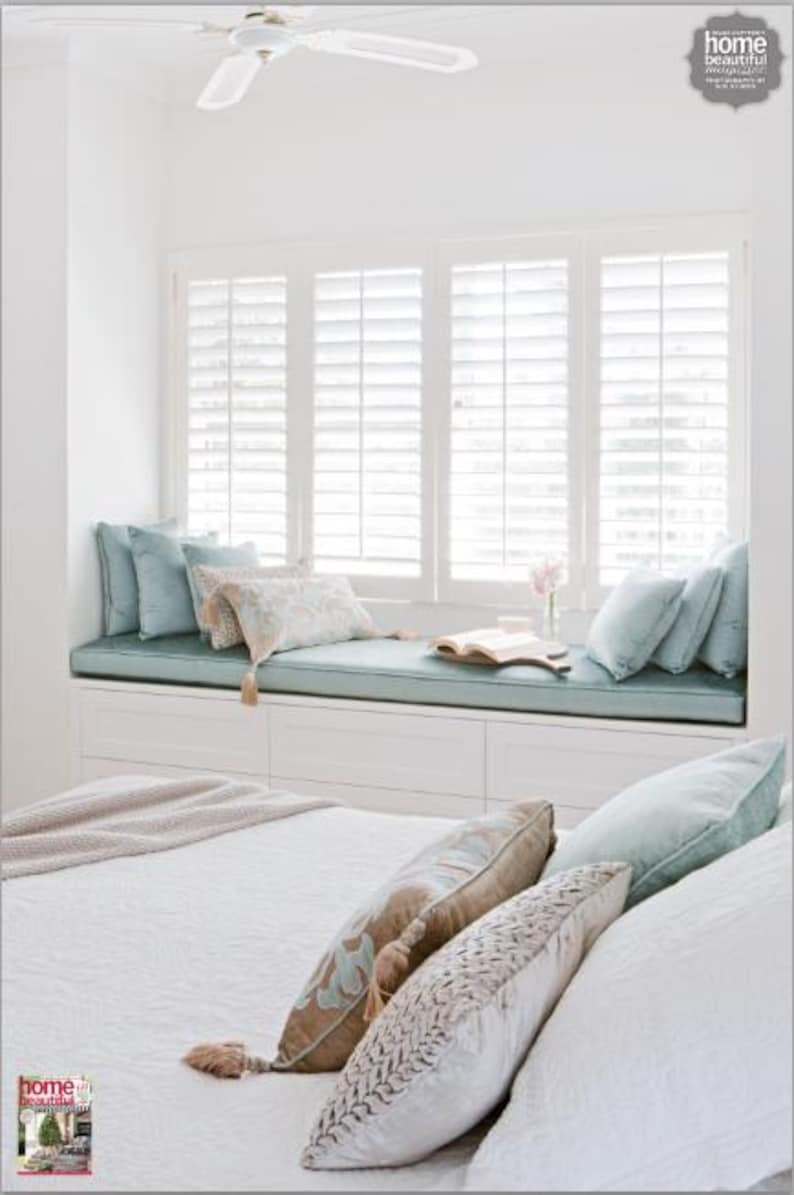May include: A bright, airy bedroom with a window seat and bed. The window seat has a light blue cushion and pillows. The bed has white bedding and decorative pillows. A ceiling fan is visible. The room is decorated in neutral tones.