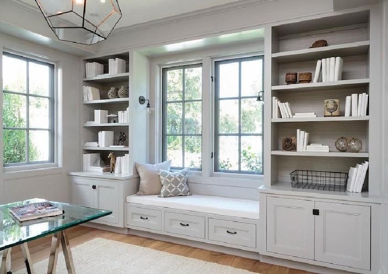 May include: A light gray home office with built-in bookshelves, a window seat, and a glass-top desk. The bookshelves are filled with white books and decorative items. The window seat has pillows. A geometric light fixture hangs from the ceiling.