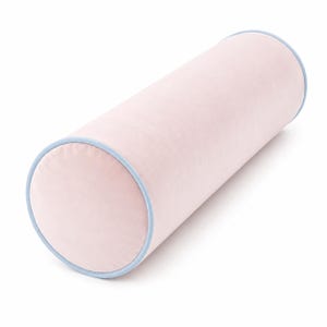May include: A cylindrical bolster pillow featuring a soft, light pink velvet fabric. The ends of the pillow are finished with a smooth, light blue piping trim that outlines the circular edges, providing a clean and defined structural appearance.