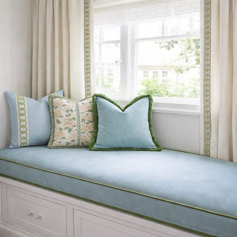 May include: A cozy window seat with light blue cushions and pillows. The pillows have green trim and decorative patterns. Cream-colored curtains frame the window, and the window seat has white storage drawers.