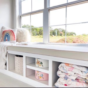 May include: A cozy window seat with a white built-in storage unit. The seat features a cream-colored cushion, a rainbow pillow, and a fluffy white throw. The storage unit includes decorative boxes and folded quilts, creating a comfortable and inviting space.