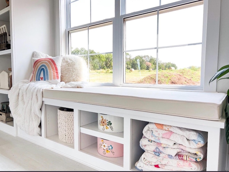 May include: A cozy window seat with a white built-in storage unit. The seat features a cream-colored cushion, a rainbow pillow, and a fluffy white throw. The storage unit includes decorative boxes and folded quilts, creating a comfortable and inviting space.