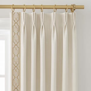 May include: Cream-colored pleated curtains with a gold-colored rod and rings. The left edge features a decorative gold geometric pattern. The curtains are hanging in front of a window, creating a soft, neutral aesthetic.