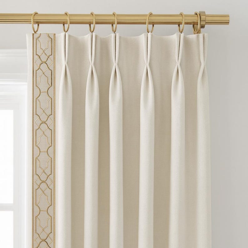 May include: Cream-colored pleated curtains with a gold-colored rod and rings. The left edge features a decorative gold geometric pattern. The curtains are hanging in front of a window, creating a soft, neutral aesthetic.