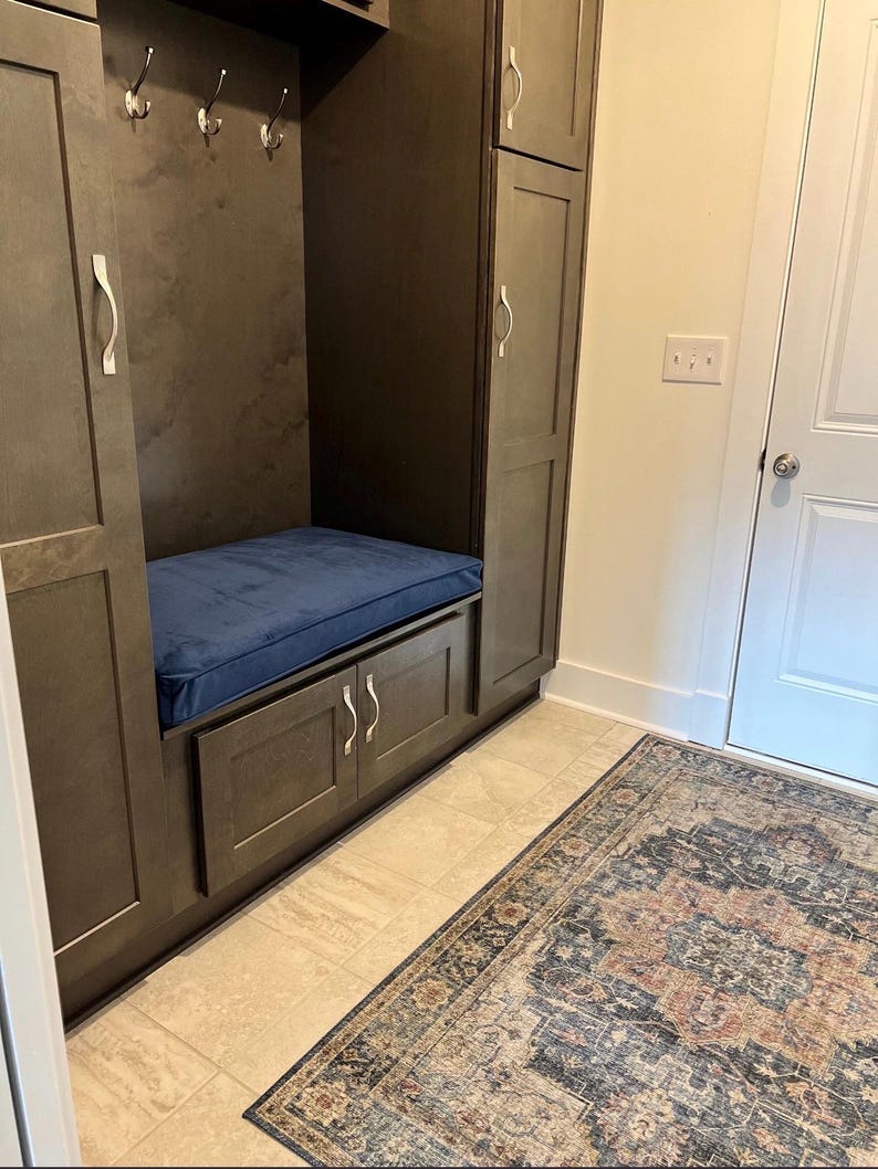 May include: A built-in entryway storage unit with dark gray cabinets, a blue cushion bench, and silver handles. The unit includes hooks for hanging items. A patterned rug lies on the floor.