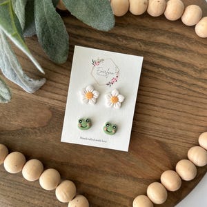 May include: A set of three pairs of stud earrings on a white card. The earrings include two pairs of white daisy earrings with yellow centers and a pair of green frog earrings. The card reads "Handcrafted with love".