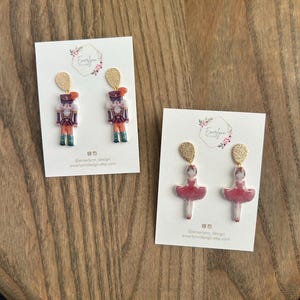 May include: Two pairs of dangle earrings on white cards. One pair features a nutcracker design with red, purple, and green details. The other pair has a ballerina design with a red tutu. Both pairs have gold-colored tops.