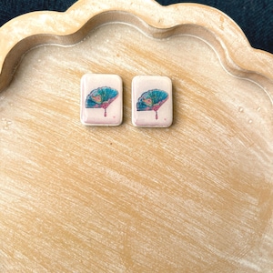May include: Two rectangular earrings with a fan design. The earrings have a light pink background with a teal and pink fan illustration. The earrings are displayed on a light brown, scalloped tray.