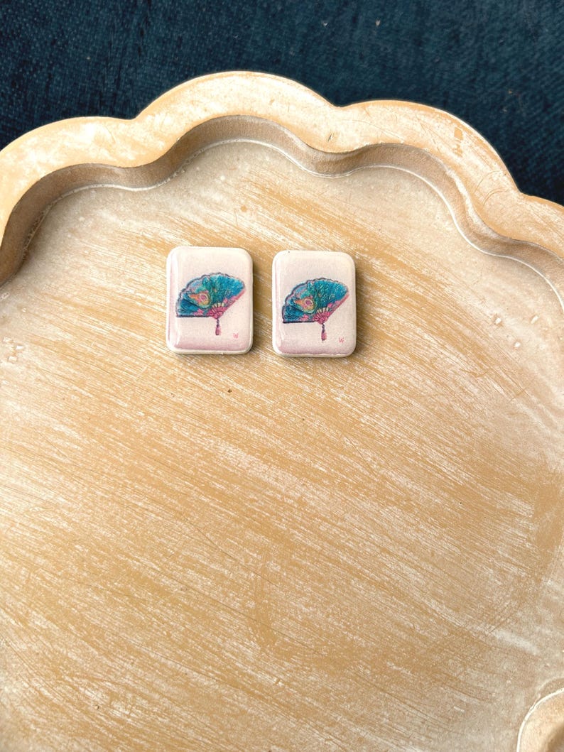 May include: Two rectangular earrings with a fan design. The earrings have a light pink background with a teal and pink fan illustration. The earrings are displayed on a light brown, scalloped tray.