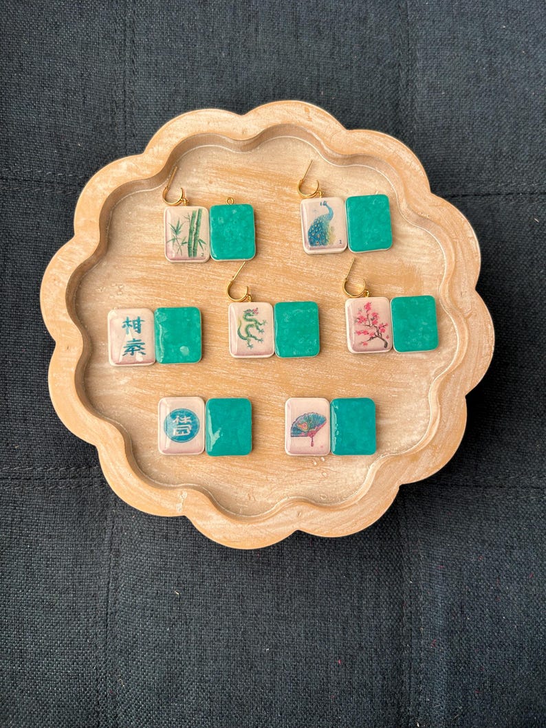 May include: A collection of square earrings with gold hooks, each featuring a unique design. The earrings have a light pink square with an image and a teal green square. Designs include bamboo, a peacock, a dragon, and a fan.