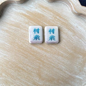 May include: Two rectangular Mahjong tiles with Chinese characters in blue on a white background. The tiles are set on a light brown, scalloped wooden tray. The tiles have a slight purple hue.