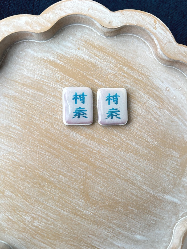 May include: Two rectangular Mahjong tiles with Chinese characters in blue on a white background. The tiles are set on a light brown, scalloped wooden tray. The tiles have a slight purple hue.