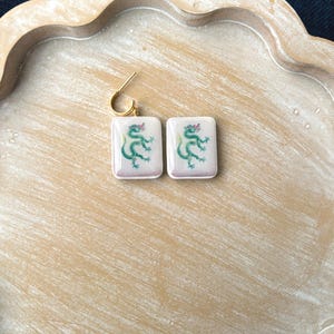 May include: A pair of square earrings with a gold-colored hook. Each earring features a white background with a green dragon design and a pink accent. The earrings are displayed on a light brown wooden tray.