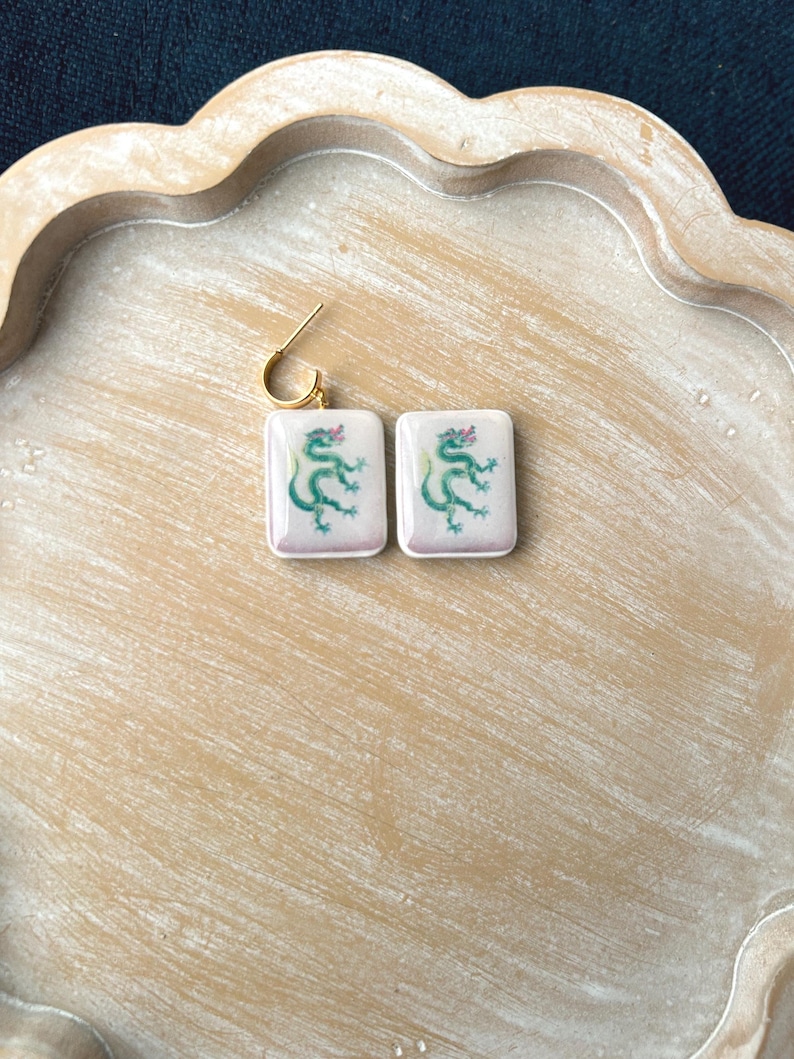 May include: A pair of square earrings with a gold-colored hook. Each earring features a white background with a green dragon design and a pink accent. The earrings are displayed on a light brown wooden tray.