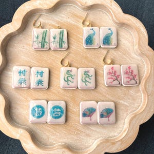 May include: A collection of square earrings with gold-colored hooks, each featuring a different design. The designs include bamboo, peacocks, Chinese characters, dragons, cherry blossoms, and fans. The earrings are displayed on a light-colored wooden tray.