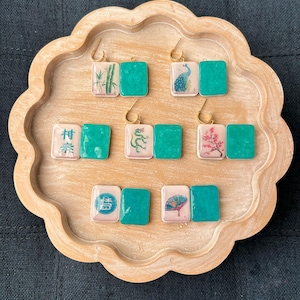 May include: A collection of handmade earrings displayed on a wooden tray. Each earring features a square tile with a gold hook. The tiles have various designs, including bamboo, a peacock, a dragon, and cherry blossoms, paired with a solid teal square.