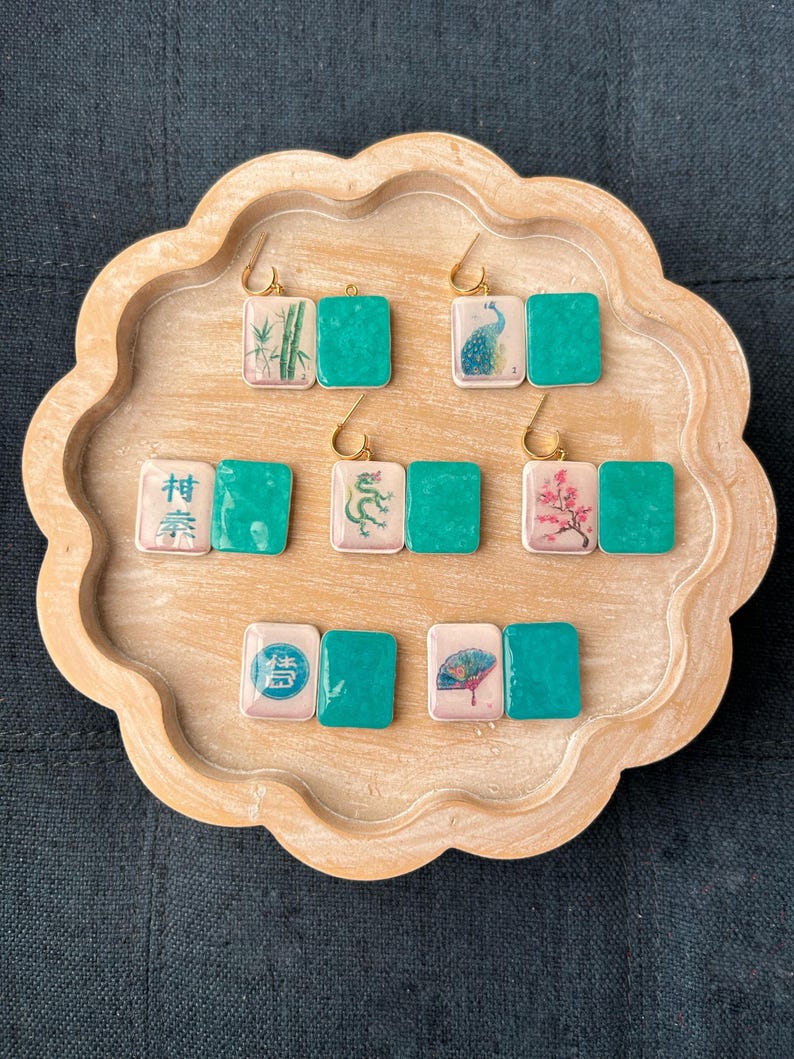 May include: A collection of handmade earrings displayed on a wooden tray. Each earring features a square tile with a gold hook. The tiles have various designs, including bamboo, a peacock, a dragon, and cherry blossoms, paired with a solid teal square.