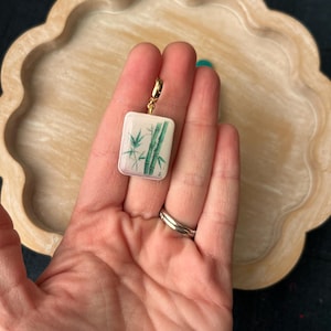 May include: A rectangular pendant with a gold-colored clasp. The pendant features a white background with a green bamboo design. The pendant is held in a hand.
