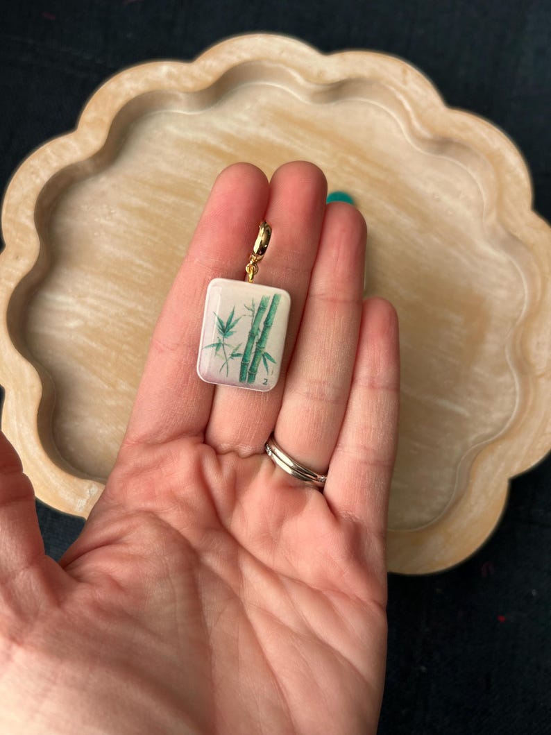 May include: A rectangular pendant with a gold-colored clasp. The pendant features a white background with a green bamboo design. The pendant is held in a hand.
