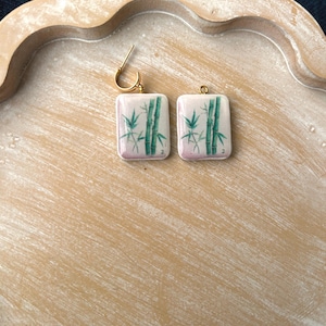 May include: A pair of square earrings with a gold-colored hoop. Each earring features a white background with a green bamboo design and a pink gradient at the bottom. The earrings are displayed on a light brown wooden tray.