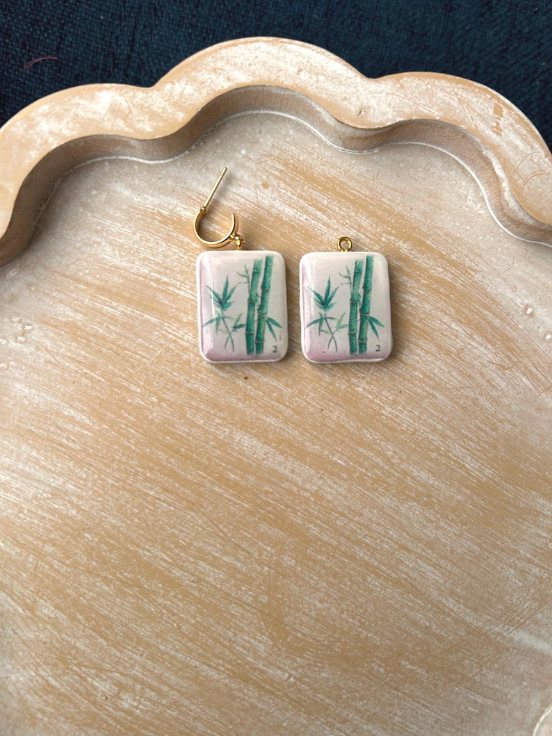 May include: A pair of square earrings with a gold-colored hoop. Each earring features a white background with a green bamboo design and a pink gradient at the bottom. The earrings are displayed on a light brown wooden tray.