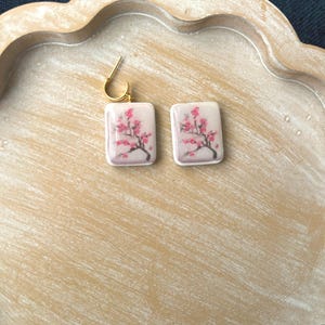 May include: A pair of square earrings with a gold-colored hoop. Each earring features a pink and red floral design on a white background. The earrings are displayed on a light brown wooden tray.