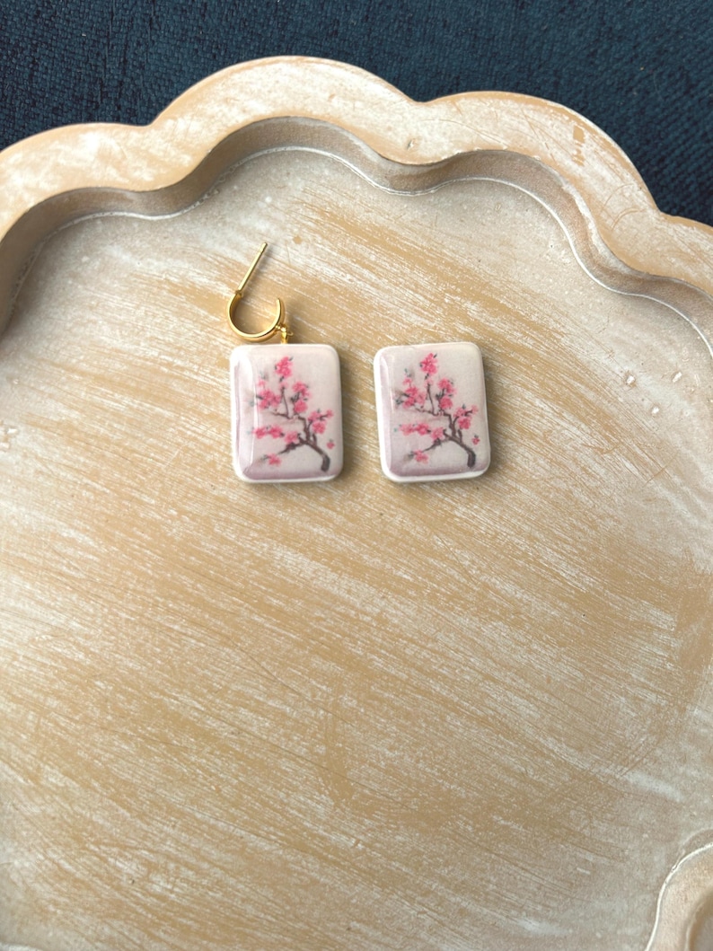 May include: A pair of square earrings with a gold-colored hoop. Each earring features a pink and red floral design on a white background. The earrings are displayed on a light brown wooden tray.