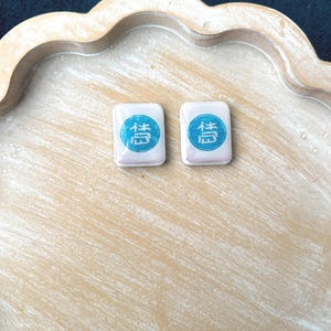 May include: Two rectangular, white Mahjong tiles with a blue and teal symbol are displayed on a light brown, scalloped tray. The tiles are side by side, and the tray has a distressed, painted finish.