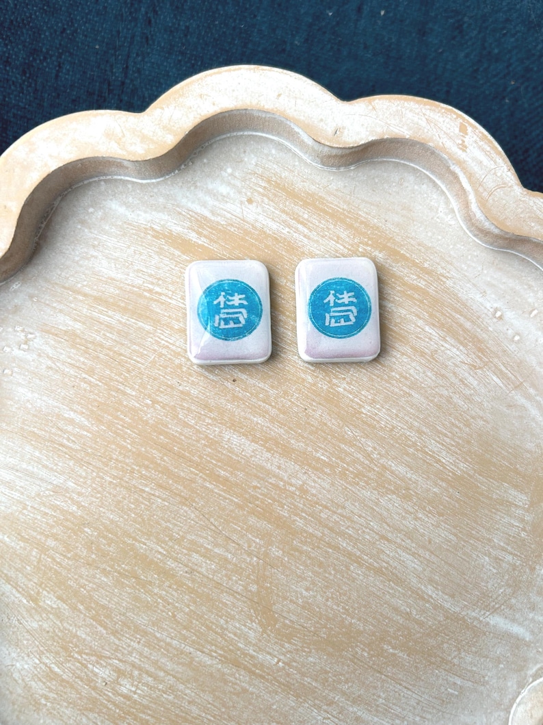 May include: Two rectangular, white Mahjong tiles with a blue and teal symbol are displayed on a light brown, scalloped tray. The tiles are side by side, and the tray has a distressed, painted finish.