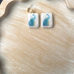 May include: A pair of square earrings with a gold-colored hoop. Each earring features a peacock design in shades of blue and green on a white background. The earrings are displayed on a light brown, scalloped tray.