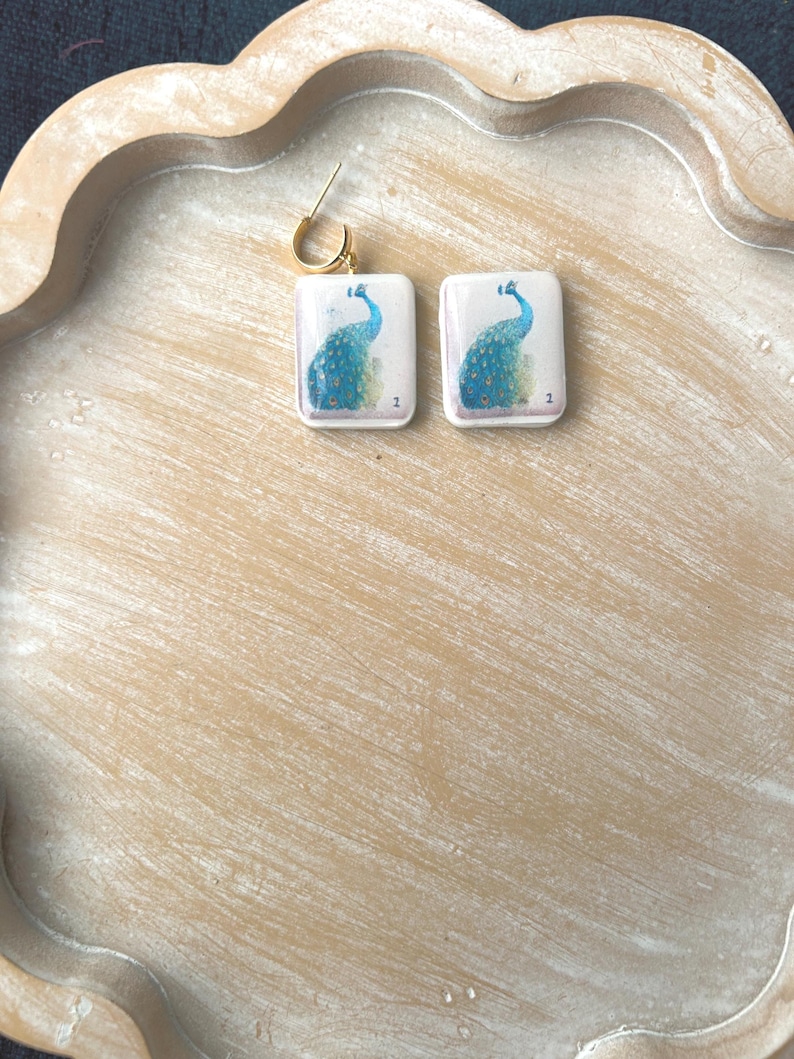 May include: A pair of square earrings with a gold-colored hoop. Each earring features a peacock design in shades of blue and green on a white background. The earrings are displayed on a light brown, scalloped tray.