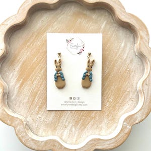 May include: A pair of dangle earrings featuring a bunny design. The earrings are attached to a white card with the text "emerlynn design" and social media handles. The earrings are on a light brown wooden tray.