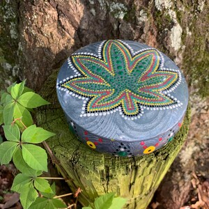 May include: A round, gray decorative box with a hand-painted cannabis leaf design in green, yellow, and red. The box is adorned with small dots and sits on a weathered wooden post, surrounded by green leaves.
