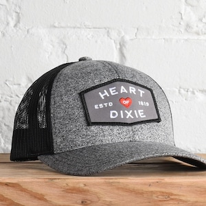 May include: A gray and black trucker hat with a black mesh back. The front of the hat has a gray patch with white text that reads "HEART OF DIXIE ESTD 1819" and a red heart.