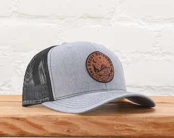 May include: A heather gray baseball cap with a black mesh back and a curved brim. The front features a circular brown leather patch with a black graphic of a fish, a crescent moon, and the text THE GREAT OUTDOORS printed in black.