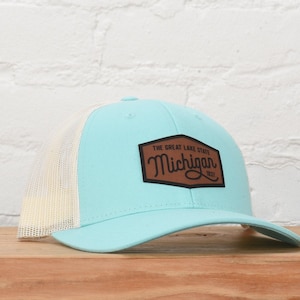 May include: A light blue and white trucker hat with a brown patch that says "The Great Lake State Michigan 1837".