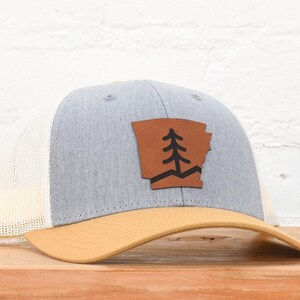 May include: A gray and beige trucker hat with a brown leather patch featuring a pine tree and the outline of the state of Arkansas.