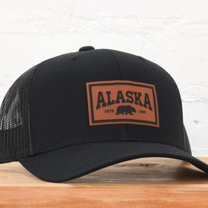May include: Black trucker hat with a brown leather patch that says "Alaska Est. 1959" and a black bear silhouette.