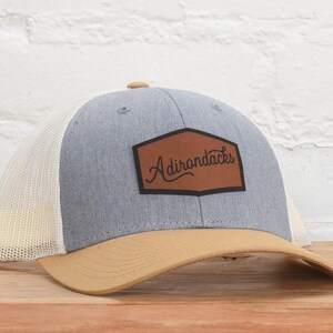 May include: A gray and tan trucker hat with a brown leather patch that says "Adirondacks" in a cursive font.