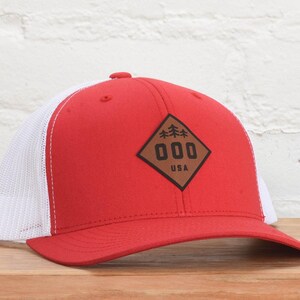 May include: Red and white trucker hat with a brown leather patch that says "000 USA" and has a small image of three trees on it.