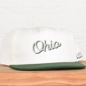May include: A white baseball cap with a green brim. The word "Ohio" is written across the front in a cursive font with a green outline. The cap has a classic, flat-brim design.