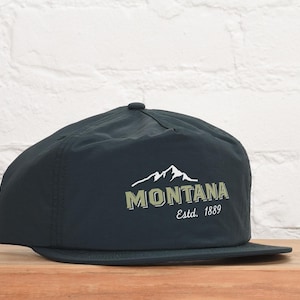 May include: A black cap with a flat brim. The cap features the word "MONTANA" in a green and white design, with a mountain graphic above the text. The text "Est. 1889" is below the word "MONTANA".