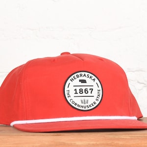May include: A red flat-brimmed cap with a white rope detail along the brim. The cap features a circular patch with the text "NEBRASKA 1867 THE CORNHUSKER STATE" and a state outline.