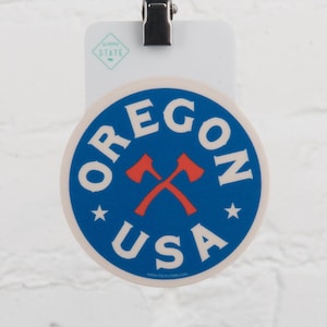 May include: A round sticker with a blue background and white text that reads "OREGON USA." Two crossed red axes are in the center. Two white stars are on either side of the axes. The sticker is held by a clip.