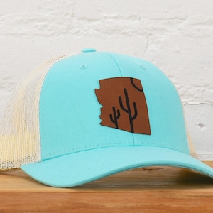 May include: A light blue and beige trucker hat with a brown leather patch featuring a silhouette of the state of Arizona with a cactus design.