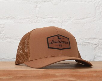 May include: A brown trucker hat featuring a mesh back and a solid front panel. A black hexagonal patch is centered on the front, displaying the text Louisiana, the number 1812, and a small silhouette of the state of Louisiana.