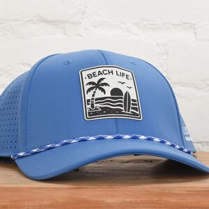May include: A blue baseball cap with a perforated side panel and a white braided cord across the brim. A rectangular patch on the front features a black graphic of a palm tree, a surfboard, and the sun with the text BEACH LIFE printed above.