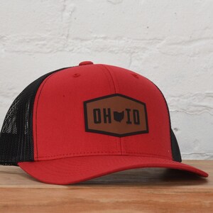 May include: Red and black trucker hat with a brown leather patch. The patch features the letters "OH IO" and an outline of the state of Ohio. The hat is sitting on a wooden surface.